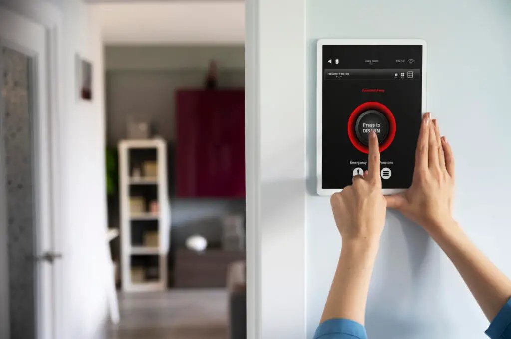 A person’s hand is pressing a red button labeled “Press to Disarm” on a wall-mounted touchscreen security system in a modern home. The background shows a bookshelf and other furniture in a well-lit room.