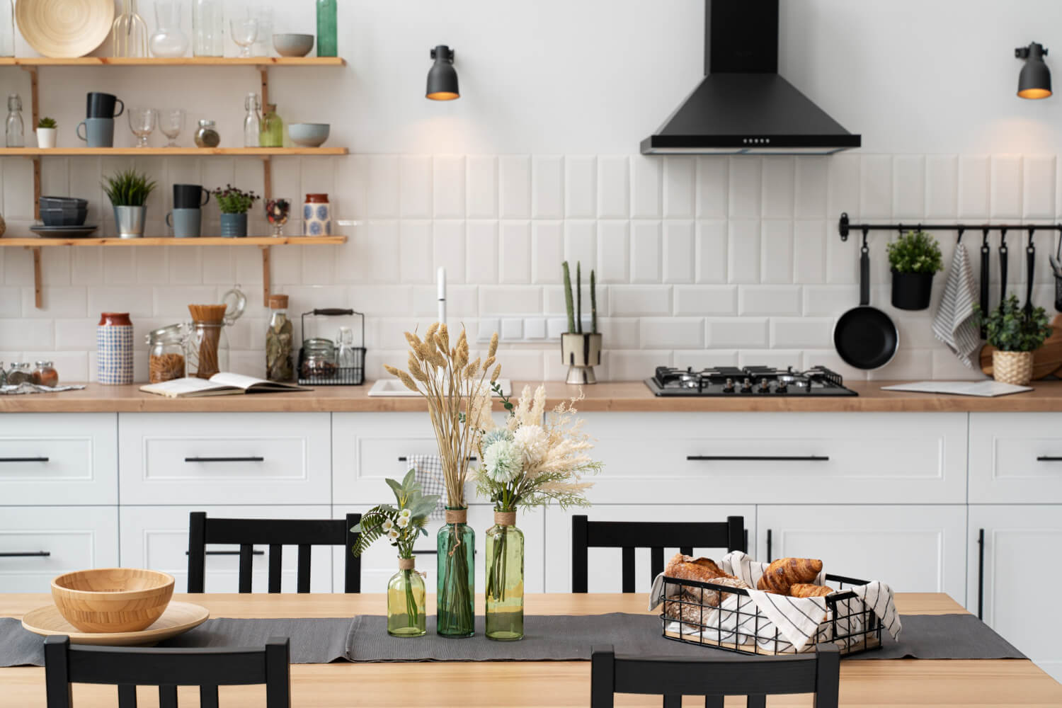 A modern kitchen with white cabinets and wooden countertops, open shelves with dishes and plants, and a dining table set with green glass vases, flowers, and a basket of croissants.