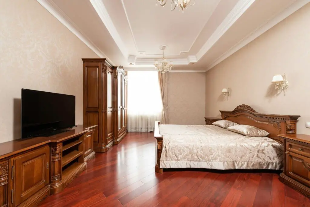 A spacious, elegant bedroom with polished wood floors, classic wooden furniture, a large bed with beige bedding, a TV on a wooden stand, and soft natural light from a window with sheer curtains.