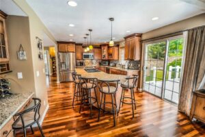 A spacious kitchen with wooden floors, brown cabinets, stainless steel appliances, and a large island with bar stools is beautifully captured in this 3d rendering, featuring pendant lights and sliding glass doors opening to a green backyard.
