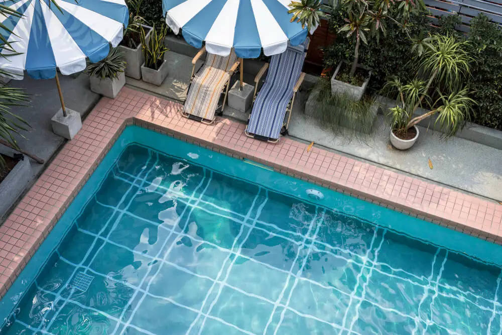 A top-down view of a swimming pool with clear blue water, surrounded by pink ceramic tiles. Two lounge chairs with blue and white striped umbrellas are poolside, and potted plants line the edge.
