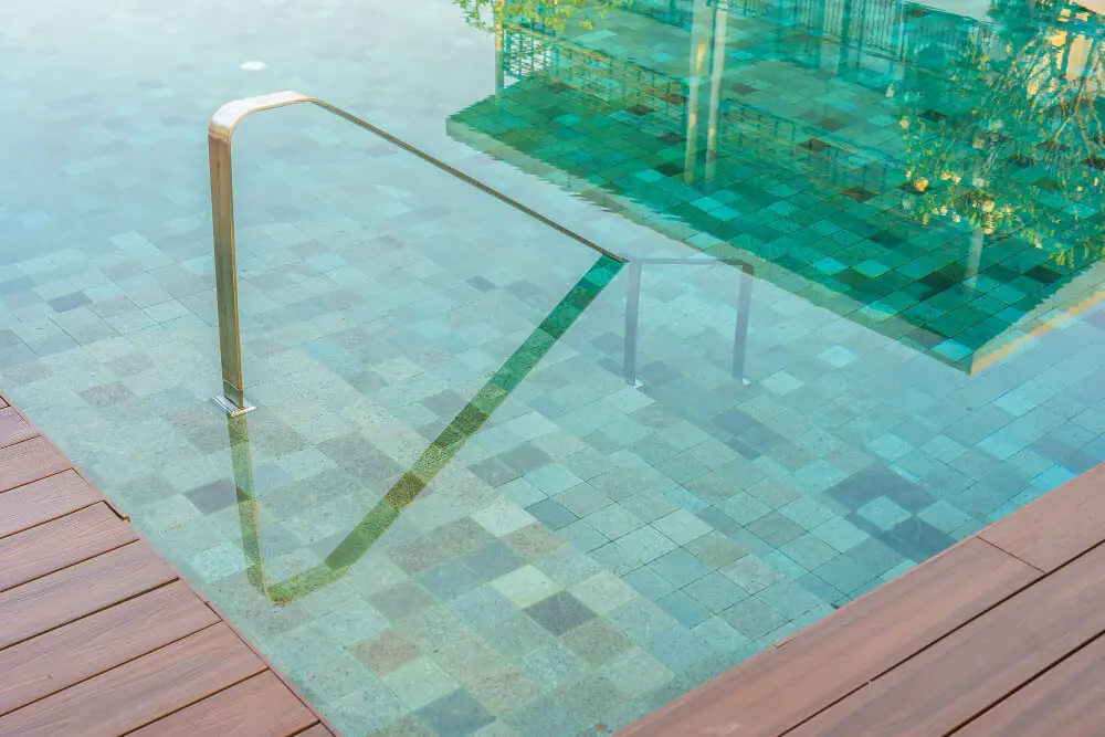 A close-up view of a swimming pool with clear turquoise water, ceramic tiles lining the floor, and a metal handrail leading into the water; wooden decking borders the pool on the left side.