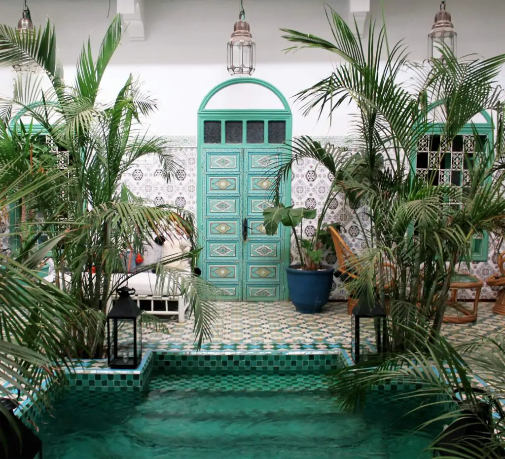 A courtyard with lush green plants, ceramic tiles, a small turquoise pool, ornate green doors, and hanging lanterns creates a vibrant, relaxing atmosphere.