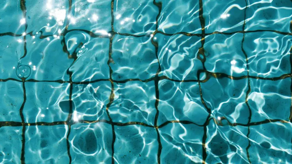 Sunlight reflects off the surface of a swimming pool, creating rippling light patterns over blue ceramic tiles underwater. The water appears clear and shimmers with wavy, bright reflections.