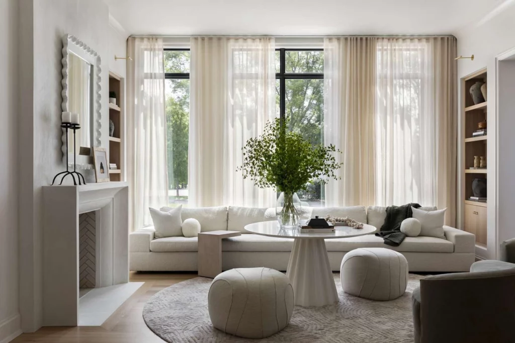 A modern small room with large windows, sheer curtains, a white sectional sofa, round coffee table, two poufs, a rug, built-in shelves, and a fireplace, decorated with greenery and neutral tones.