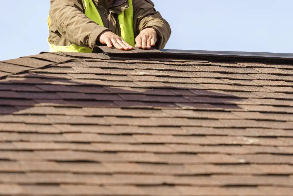 A worker wearing a jacket and safety vest uses their hands to install asphalt shingles on a rooftop under a clear sky, carefully addressing the hidden costs of roof repair during the process.