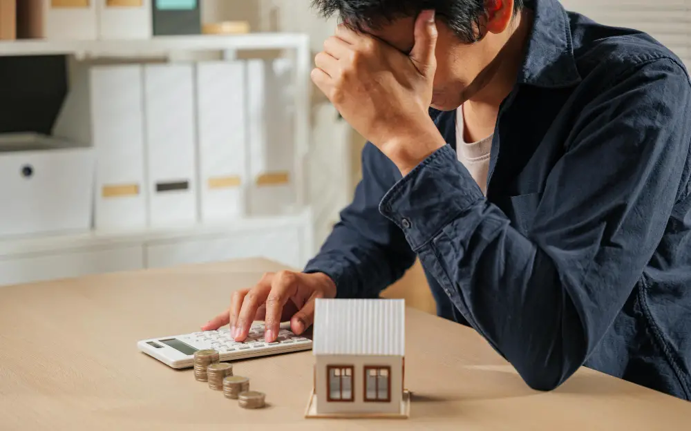 A person sits at a desk with their head in one hand, looking stressed while using a calculator. Coins and a small house model are on the table, suggesting financial concerns related to housing.