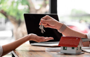 A person hands over house keys to another across a table with a small model house, a laptop, documents, and a list of 10 questions visible in the background.