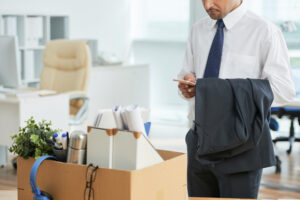 A man in business attire holds a suit jacket and checks his phone beside a box of personal belongings in an office, suggesting he is leaving—perhaps due to job relocation, which can force a sale of his home.