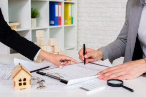 Two people in business attire sit at a desk with paperwork, model houses, and office supplies, discussing and signing documents, likely related to seller financing or a real estate contract agreement.