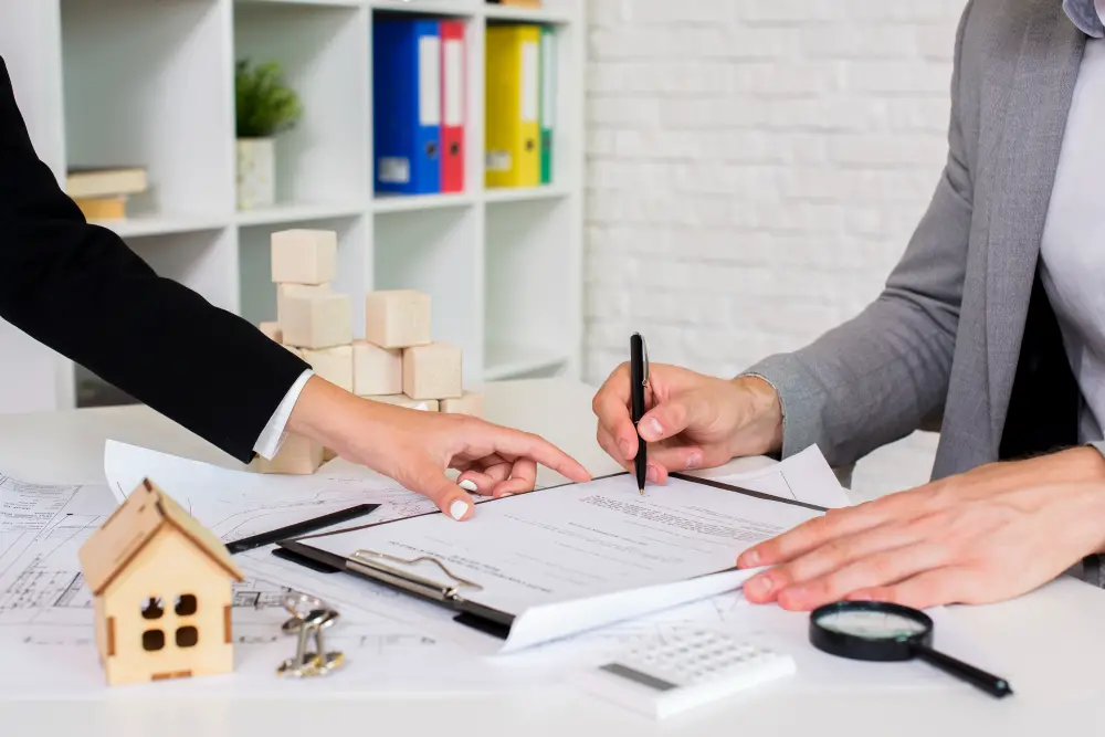 Two people in business attire sit at a desk with paperwork, model houses, and office supplies, discussing and signing documents, likely related to seller financing or a real estate contract agreement.