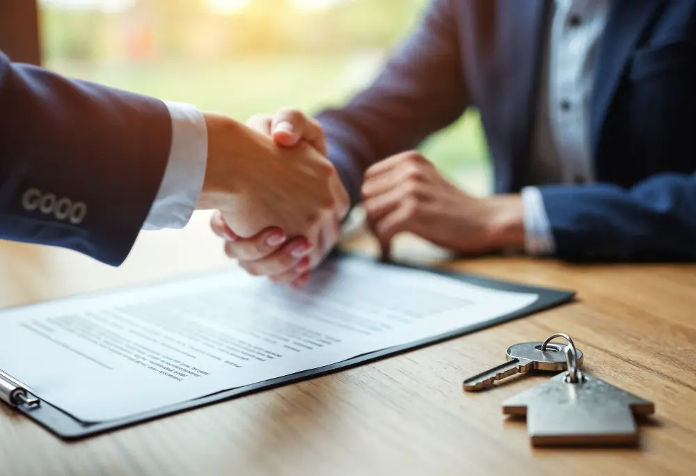 Two people in business attire shake hands across a desk with a contract and house keys on it, symbolizing a real estate or business agreement potentially involving seller financing.