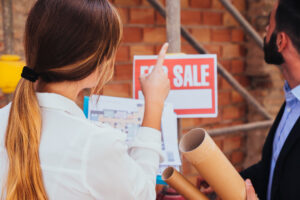 A woman and a man stand near a For Sale sign, holding rolled-up papers and looking at a building under construction with exposed brick and scaffolding.