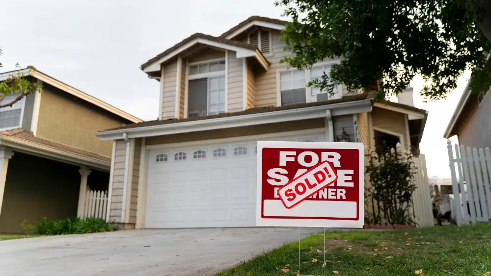 A two-story house with beige siding and a white garage door has a For Sale sign in the front yard, with SOLD! written across it in red letters.