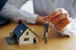 A person’s hand holds a set of house keys next to a small model house on a wooden table, symbolizing buying or renting a home.