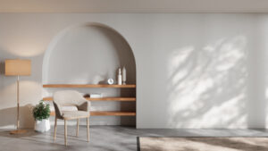 A minimalist living space with a beige armchair, floor lamp, potted plant, and arched wall niche featuring wooden shelves shows how to make your living space feel more high-end without a full renovation. Sunlight casts soft shadows on the white wall.