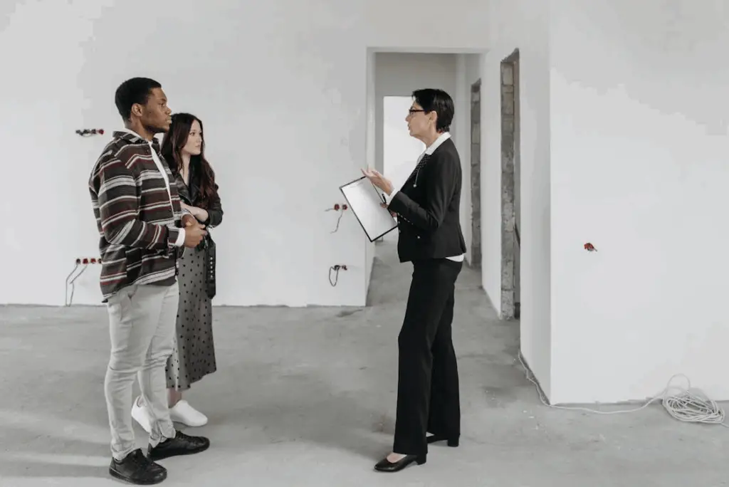 A real estate agent holding a clipboard speaks to a man and a woman inside an empty, unfinished room with white walls and exposed wires.